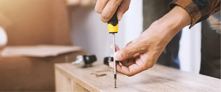 Hände drehen Schraube in Holzplatte ein, daneben Werkzeug und Bauteile auf hellem Untergrund in einer Montagearbeit.